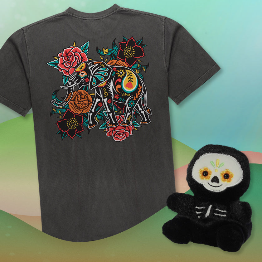 Gray t-shirt with colorful elephant design on a gradient background, next to a small sugar skull plush toy.